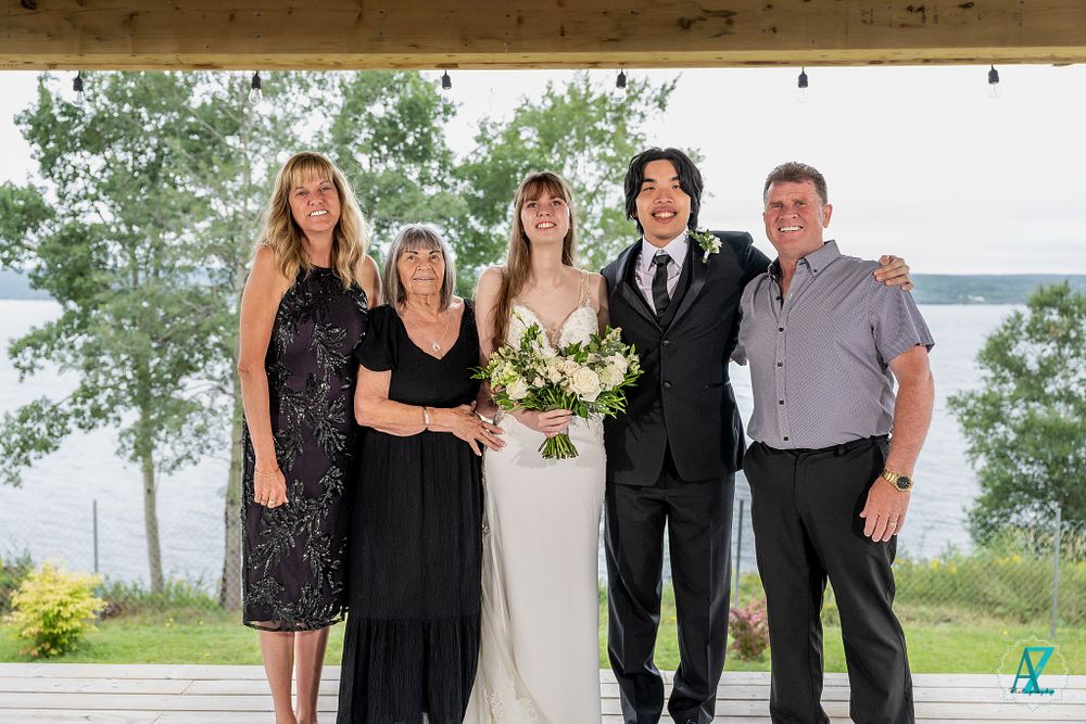 Wedding party group photo under wooden pergola with lakeside scenic backdrop.