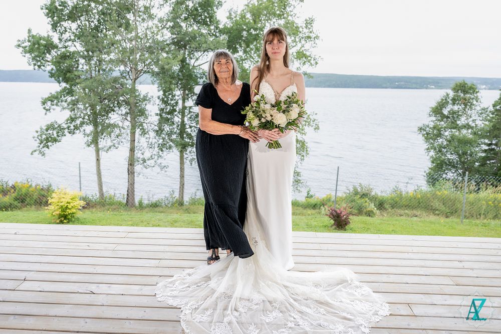 Two wedding attendees pose on wooden deck with flowing white dress and lake view.