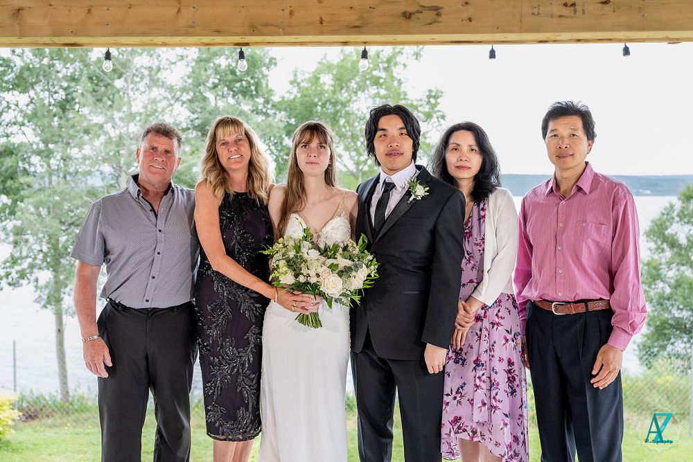 Formal group portrait of wedding party members standing together on a deck overlooking a lake.
