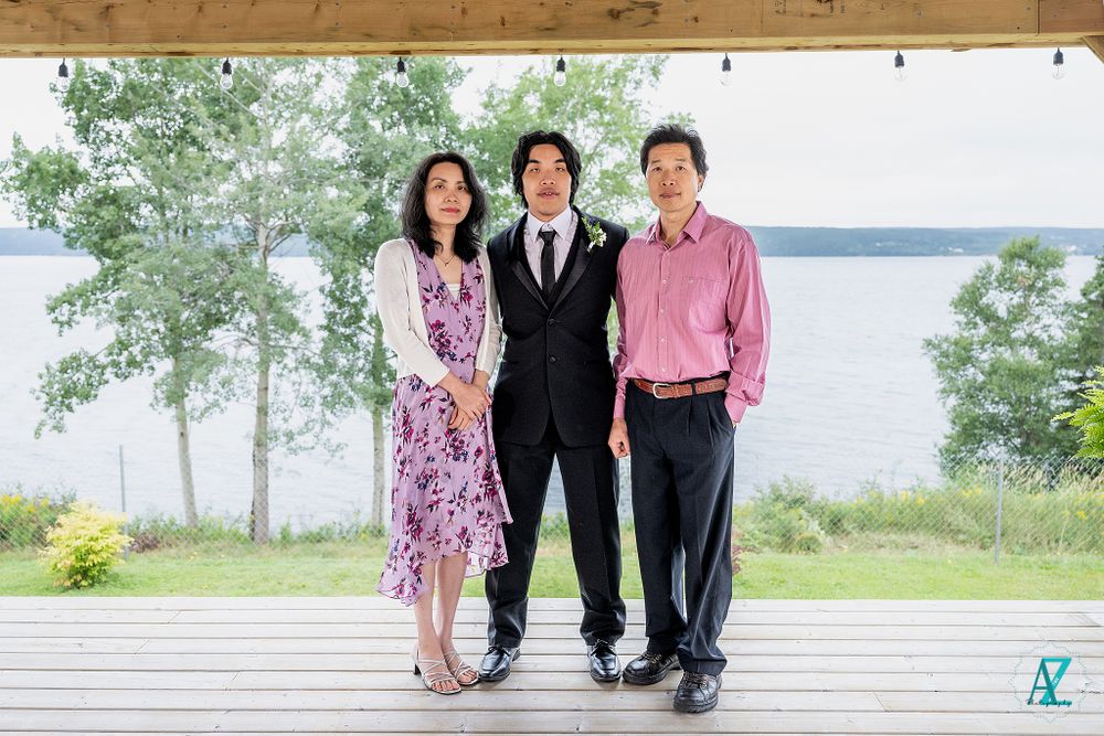 Three formally dressed guests pose on an outdoor deck with a scenic lake view in the background.