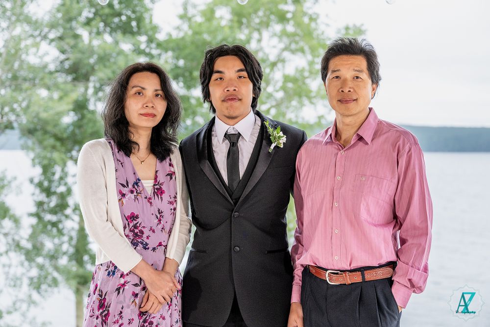 Three guests in formal attire pose at outdoor wedding venue with water and trees in background.