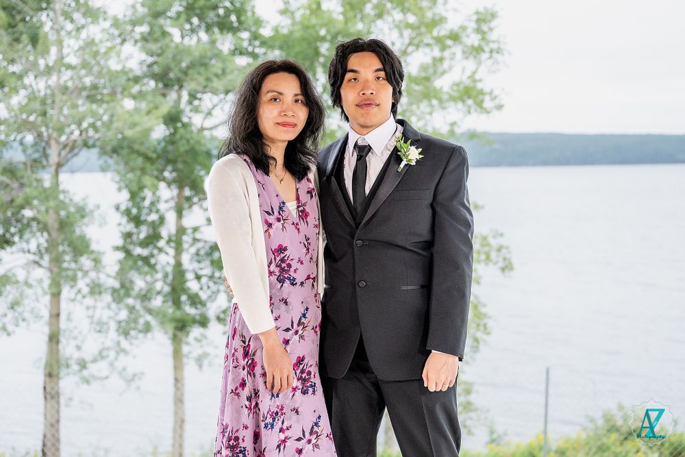 Two wedding guests pose together on deck with scenic lake and forest backdrop.