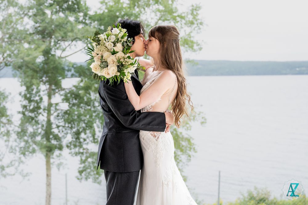 A couple shares their first kiss as newlyweds with a waterfront view in the background.