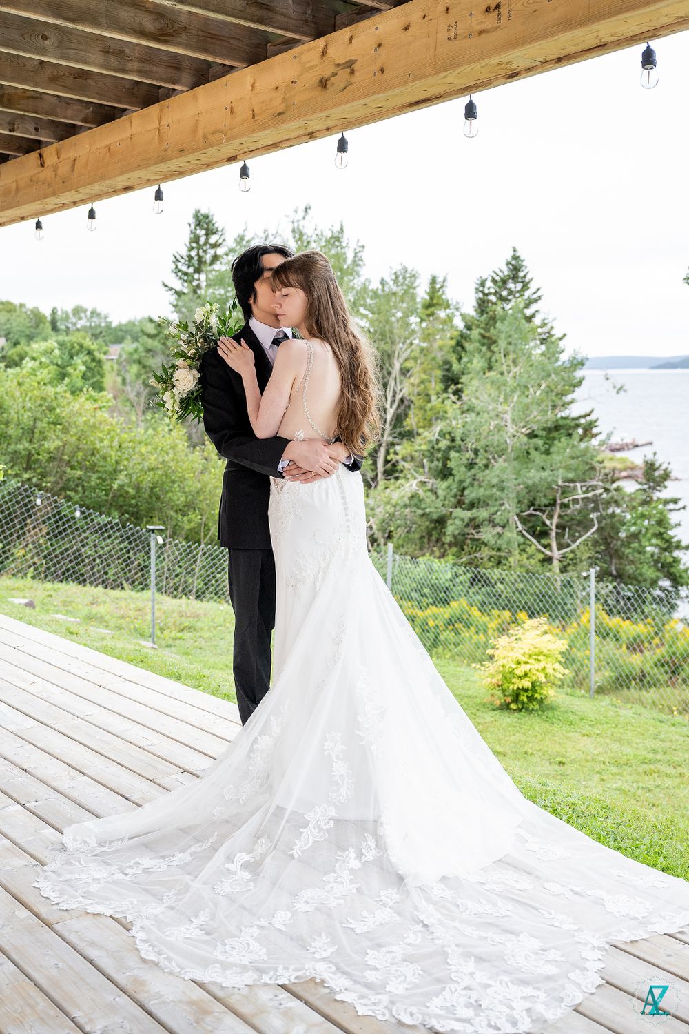 A full-length wedding portrait on a wooden deck with a flowing lace train and water view.