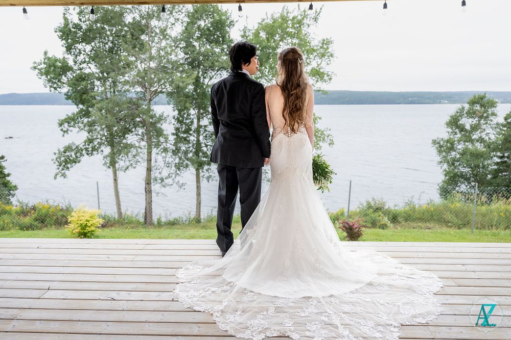 Bride and groom standing on wooden deck overlooking lake with long lace wedding dress train.