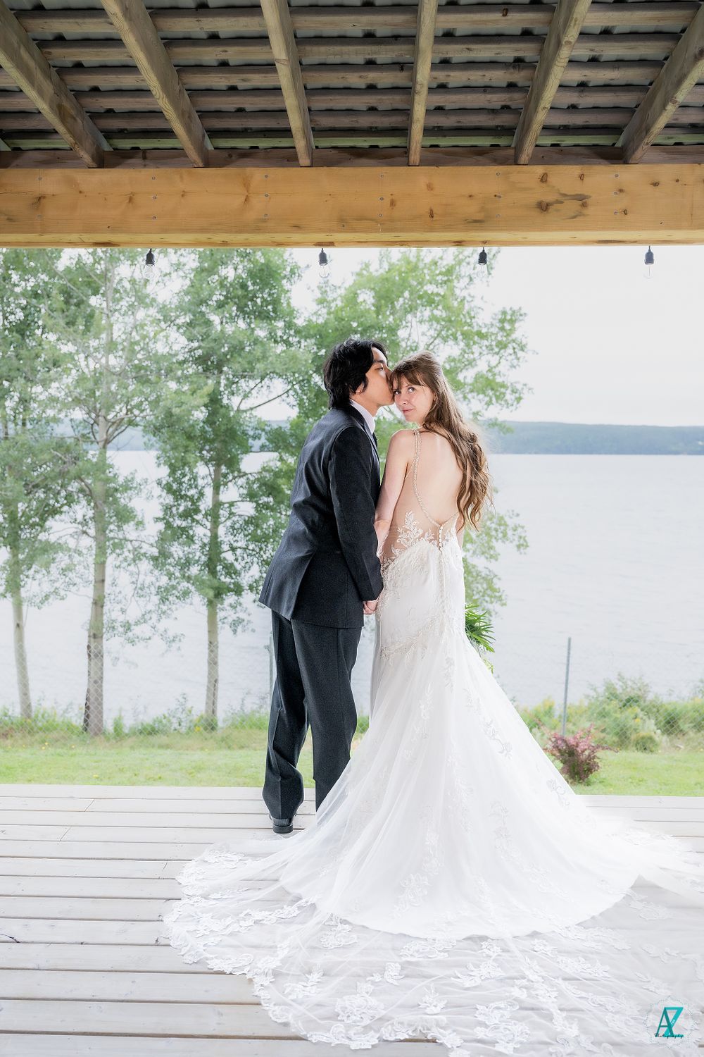 Romantic wedding couple embracing on lakeside deck with dramatic train of white lace gown.