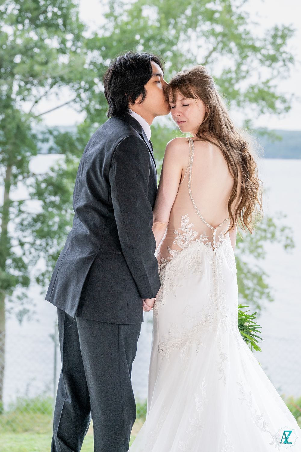 Elegant wedding photo showcasing bride's detailed backless gown as couple embraces by lake.