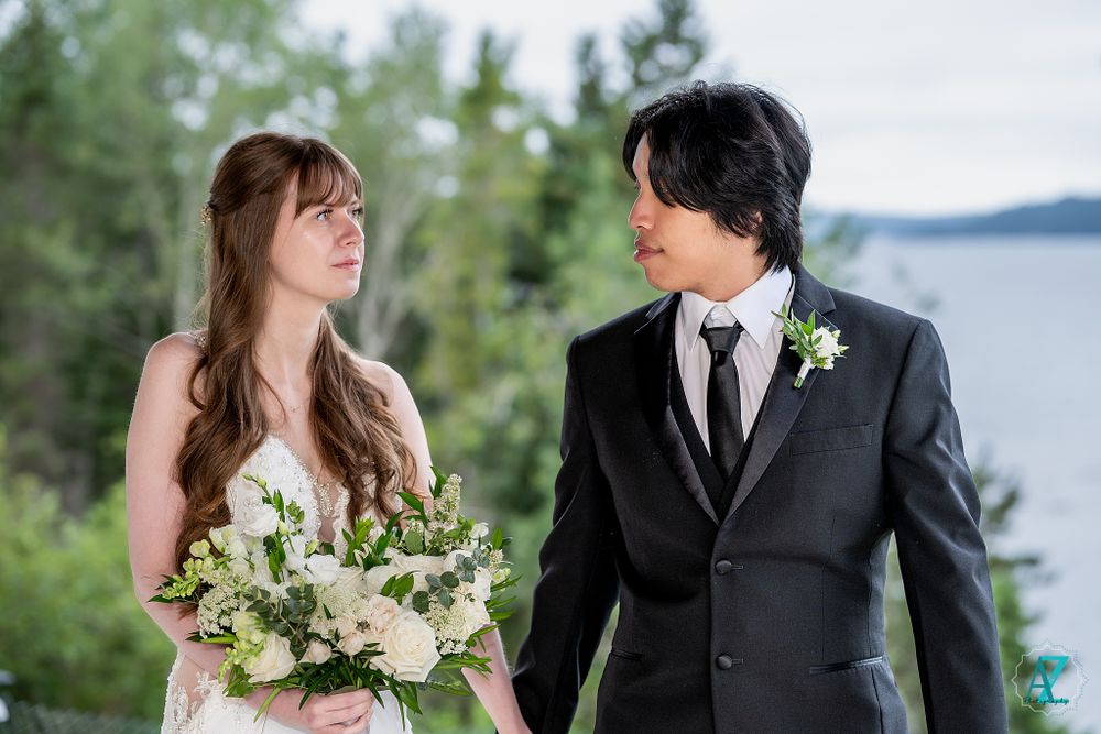 A bride in white holds a bouquet while sharing a tender moment with her groom on their wedding day by a lake.
