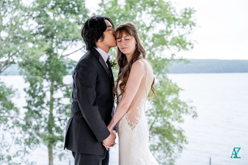 Newlyweds share an intimate moment by a serene lake with trees in the background.