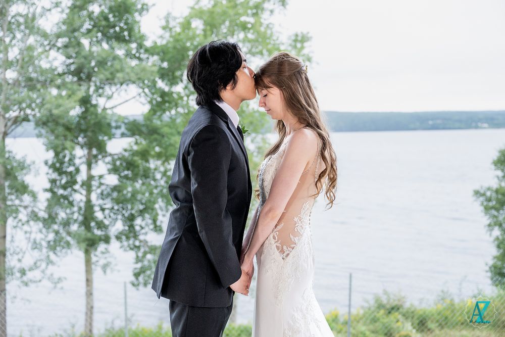 A romantic wedding portrait of a couple embracing against a lakeside backdrop with trees.