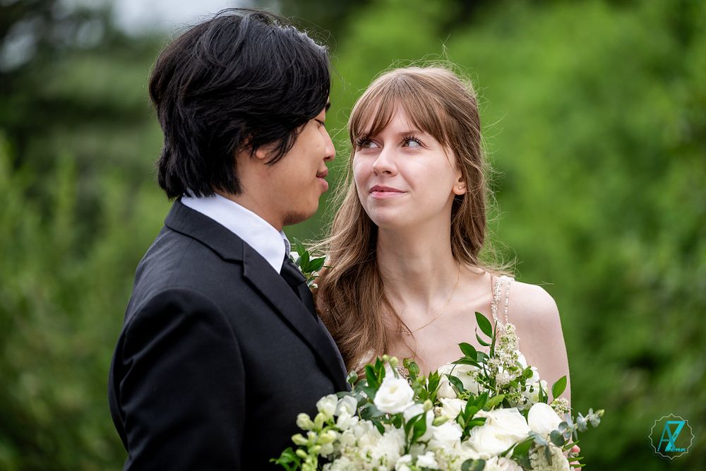 Wedding couple shares a loving look while holding a white and green floral bouquet outdoors.