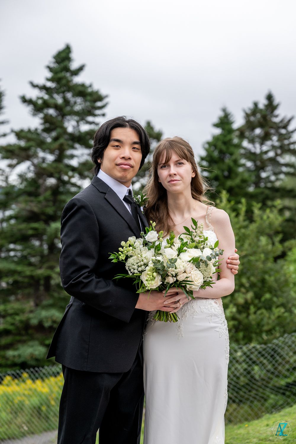 Elegant wedding portrait with couple holding white bouquet against pine forest backdrop.