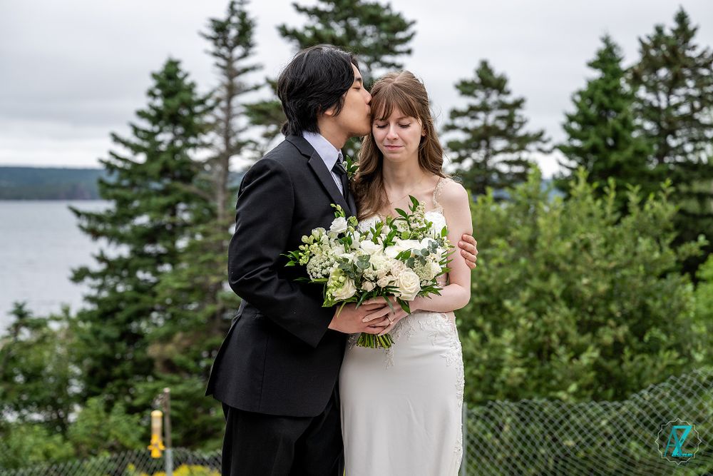 Wedding couple poses against evergreen trees and lake backdrop with elegant white bouquet.