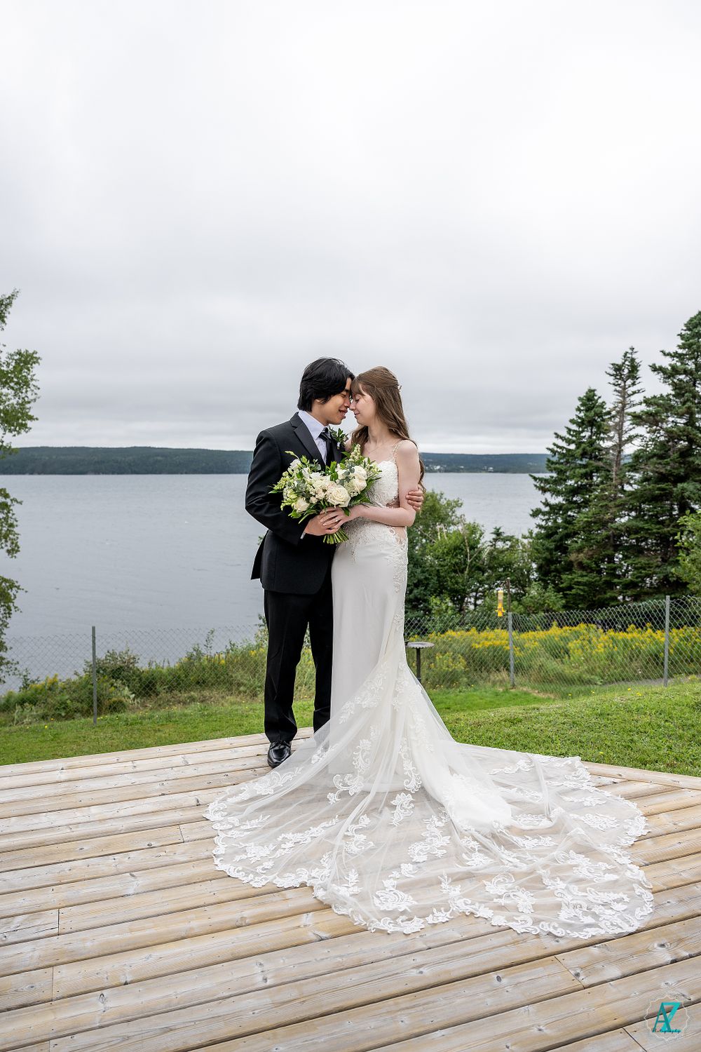 Bride in lace train gown and groom share moment on wooden deck overlooking lake view.