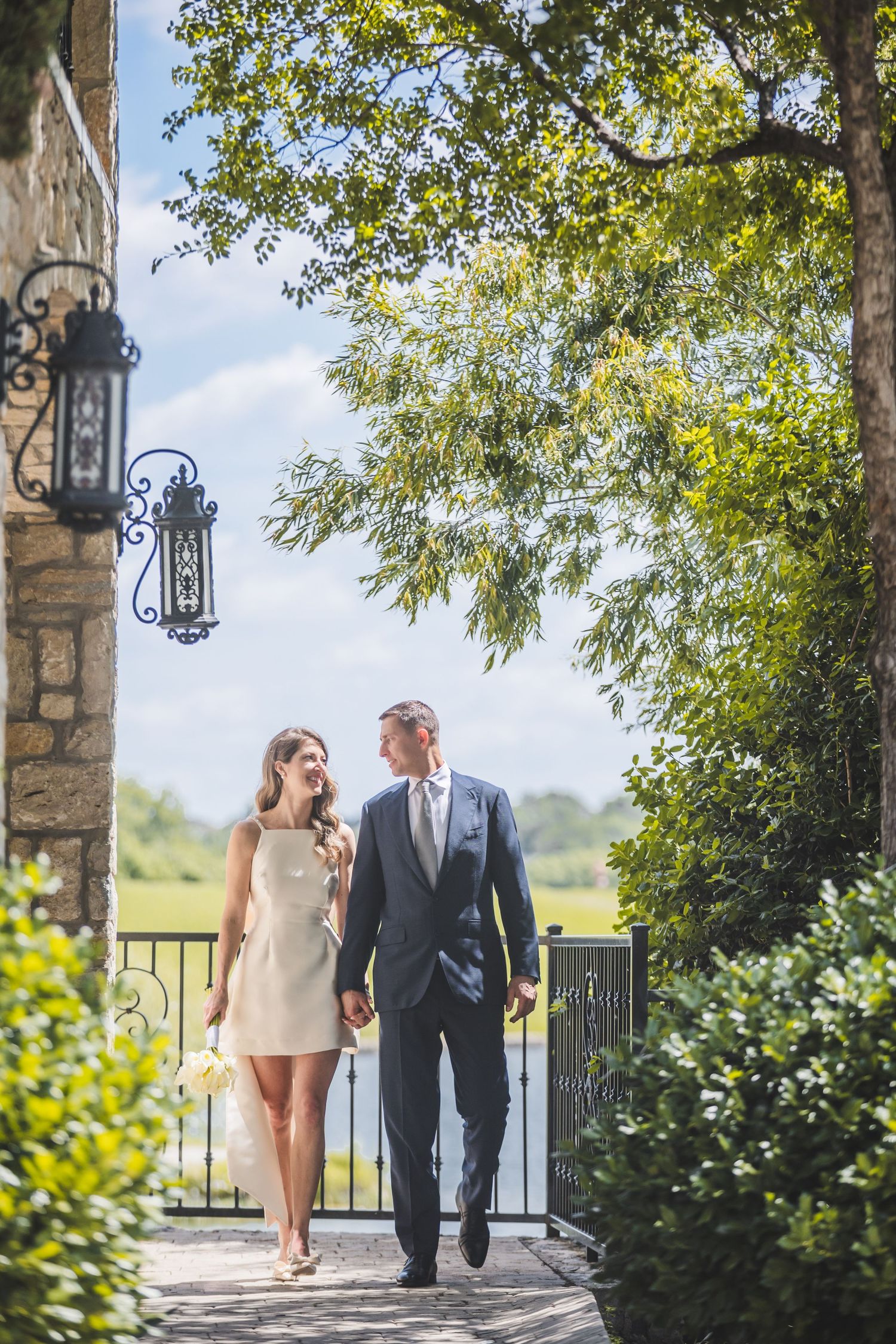A couple walks together along a stone pathway framed by lush greenery and ornate lanterns on a sunny day.