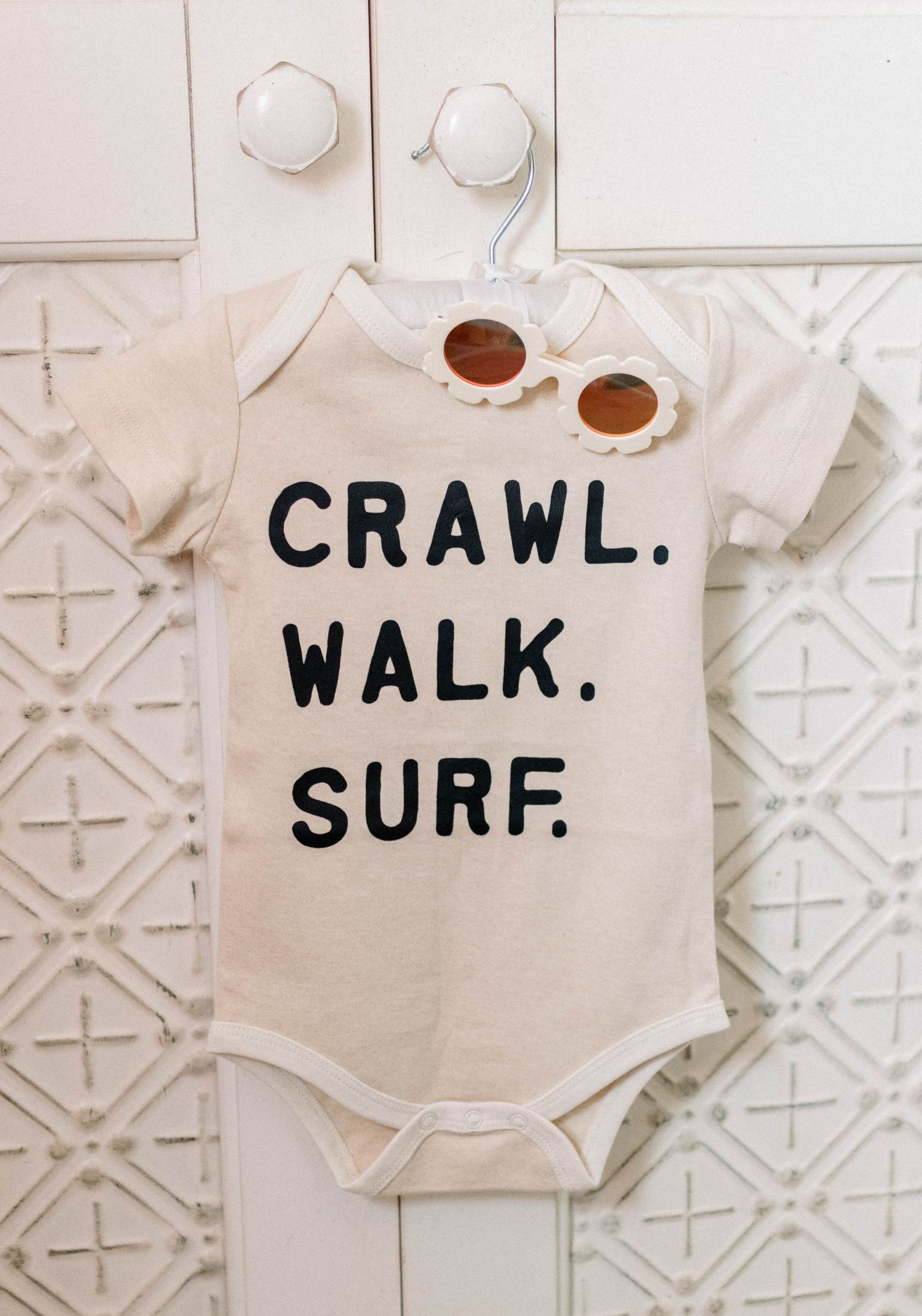 Baby onesie with text 'Crawl Walk Surf' and sunglasses hanging on white wall.
