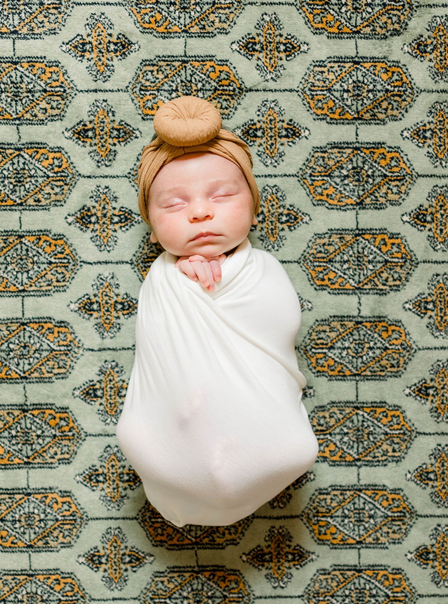Sleeping newborn in white swaddle on patterned rug with matching headpiece.