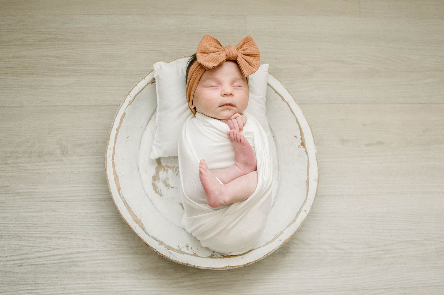 Newborn baby wrapped in white swaddle lying in round white bowl wearing peach bow.