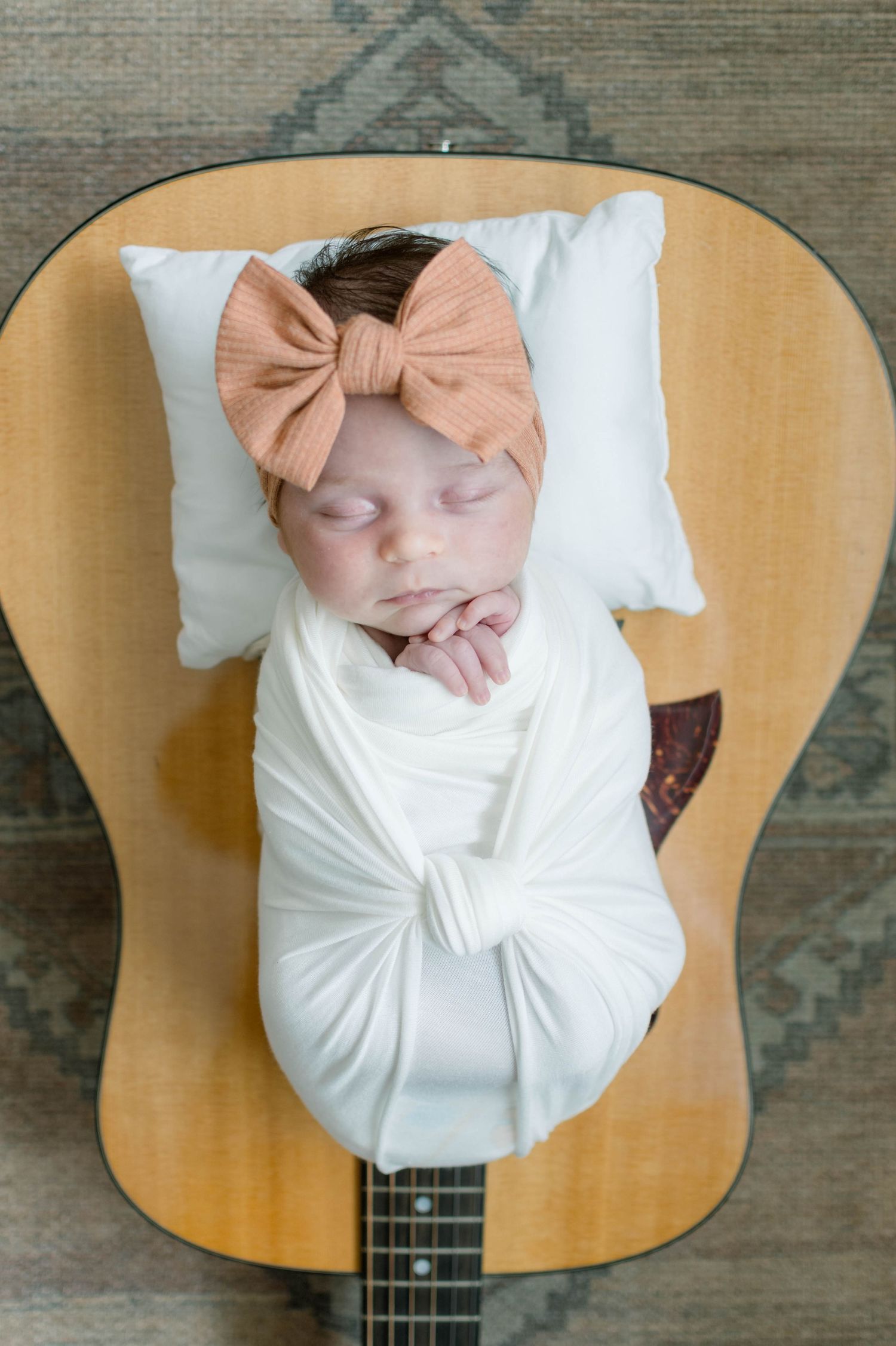 Newborn wrapped in white swaddle with peach bow headband lying on acoustic guitar.