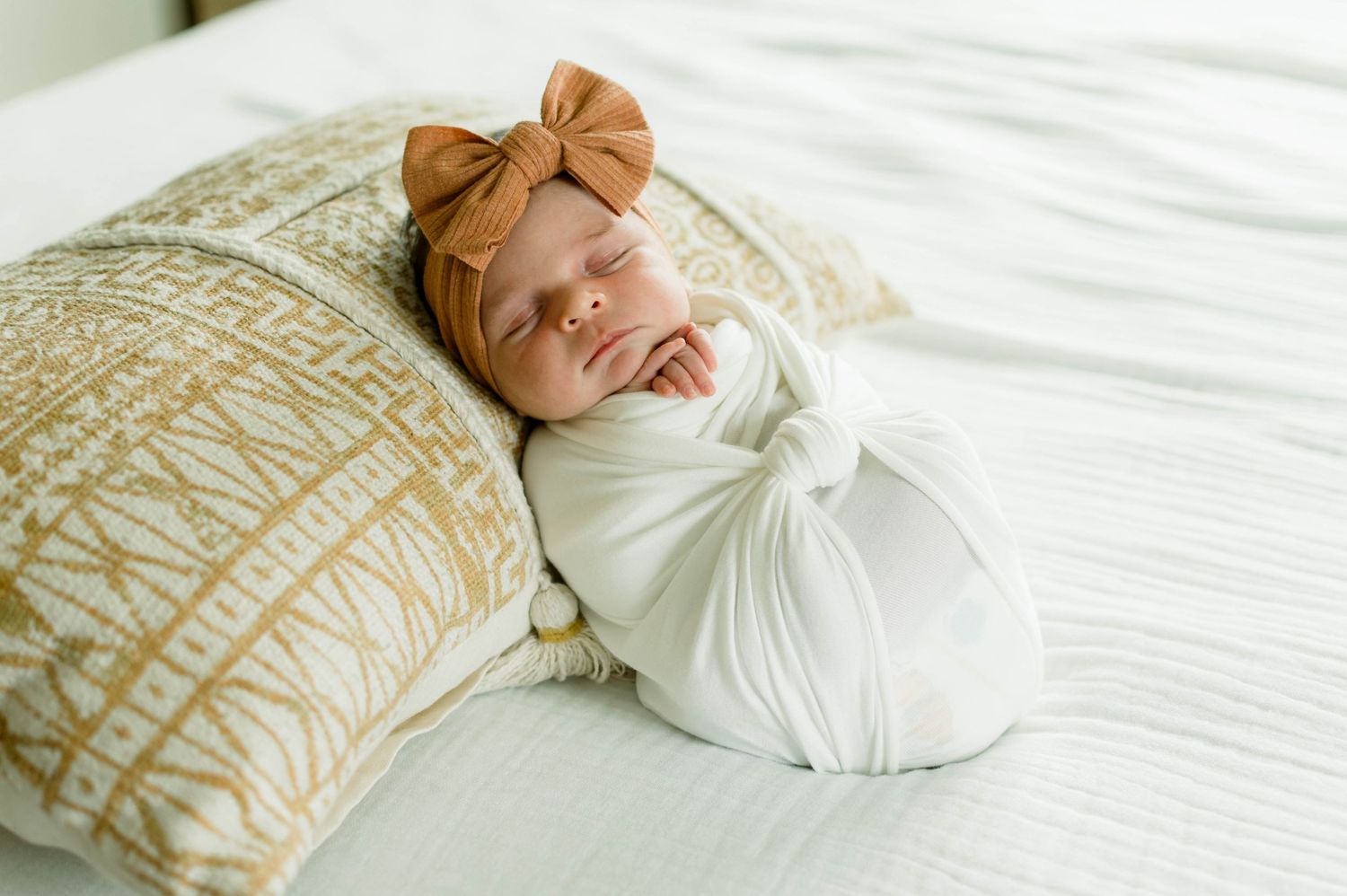 Newborn baby in white swaddle with peach bow accessory on textured bedding.