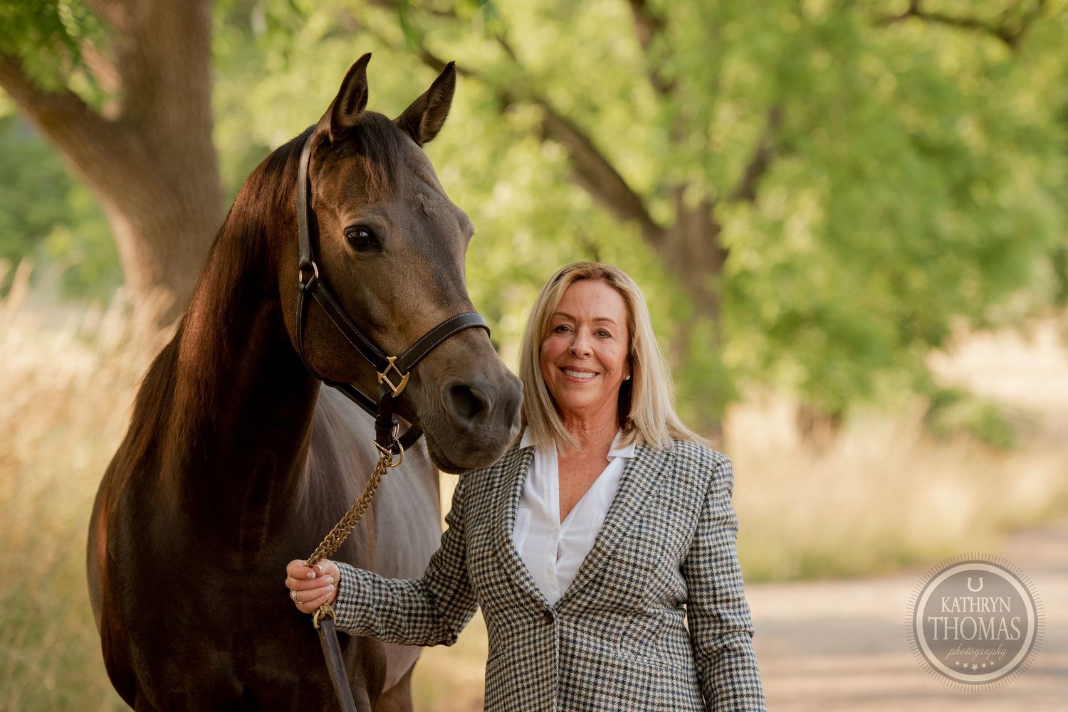 Elegant dark horse and handler in tweed blazer pose together on a country path.