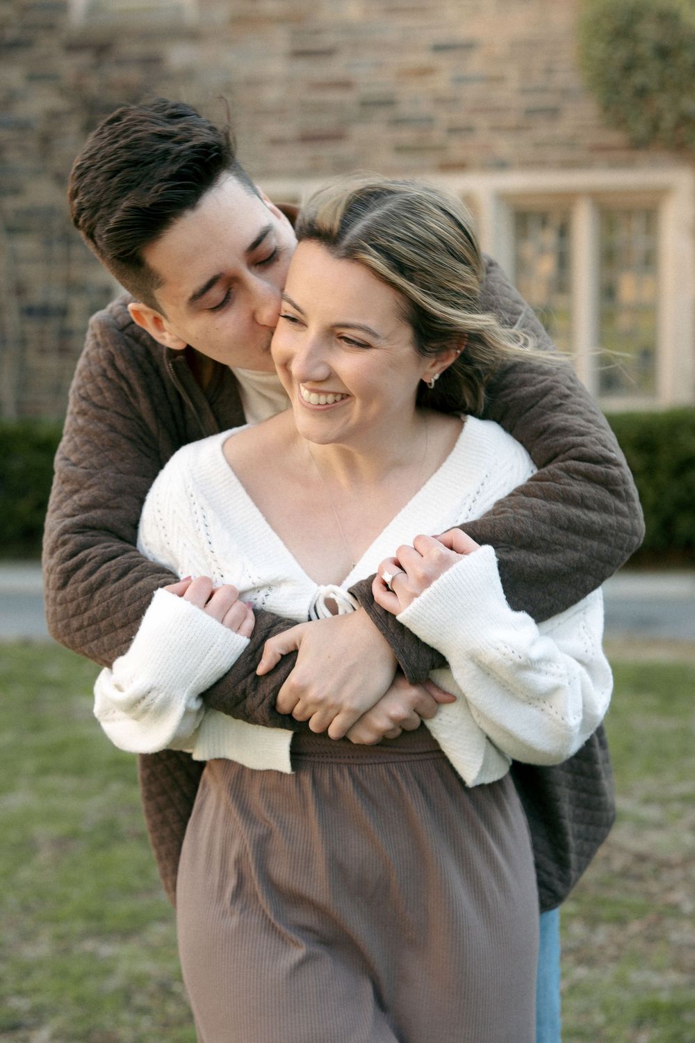 A happy couple embraces outdoors, man kissing woman on cheek while she smiles, wearing cozy sweaters near a stone building.