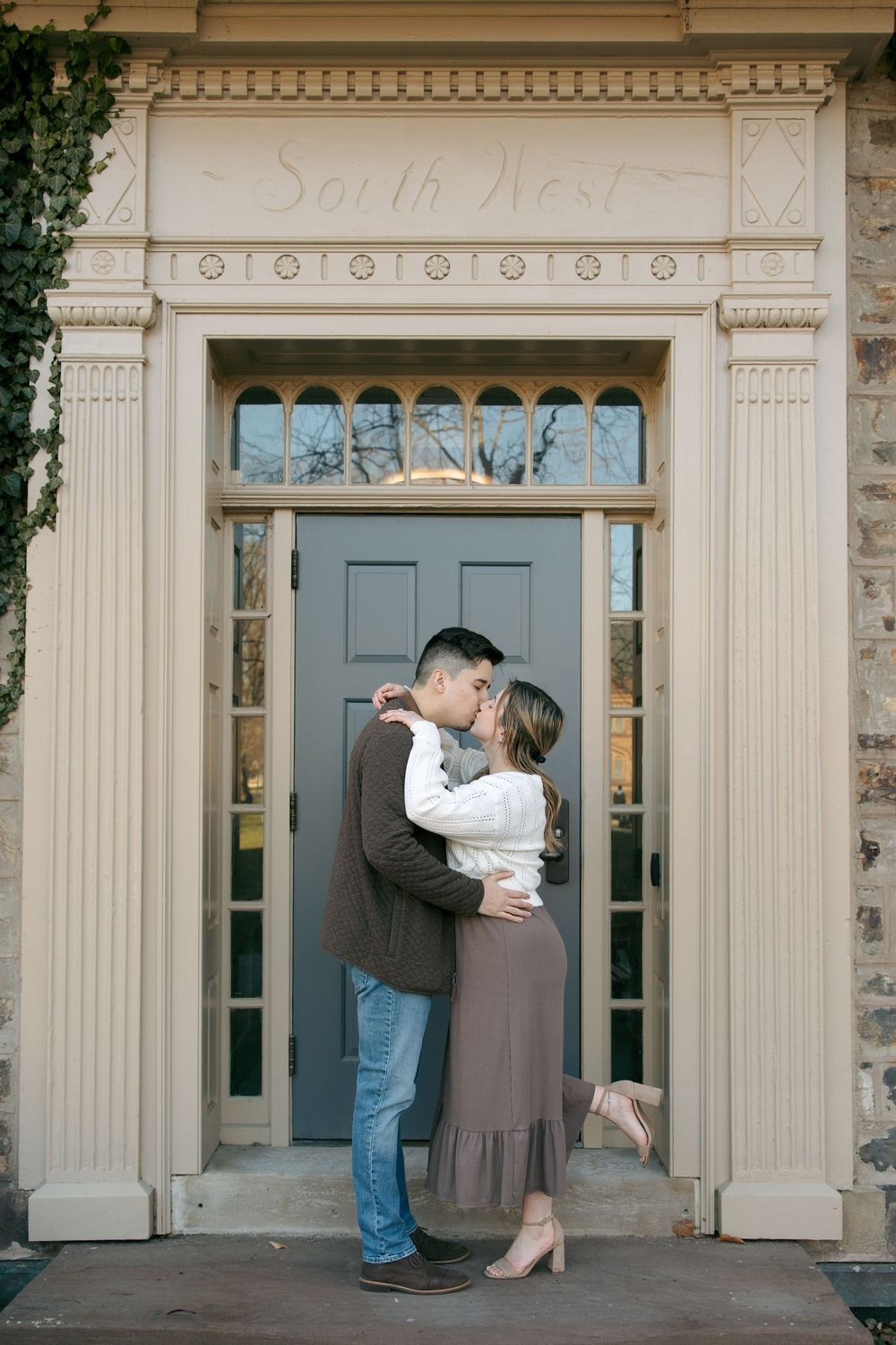 Couple sharing a romantic kiss in front of an elegant South West building entrance with ornate white trim and blue door.