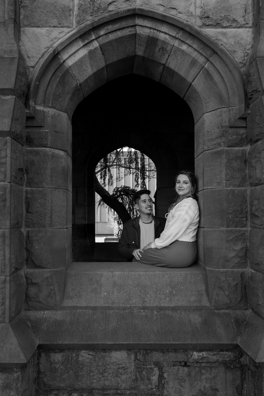 Black and white photo of a couple sitting in a gothic stone arch window of a historic building.