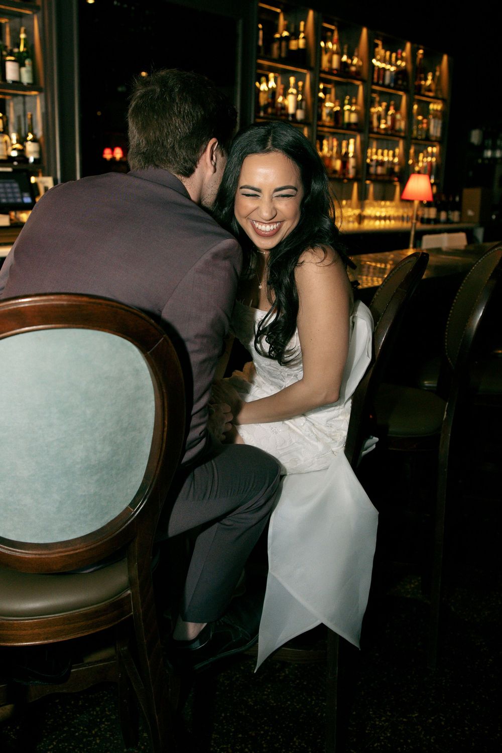 A laughing bride in a white dress sits with a groom at a dimly lit bar, sharing a joyful intimate moment.