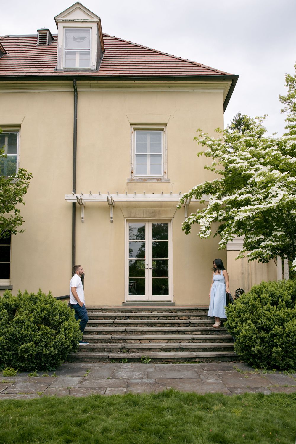 Couple standing apart on steps outside a yellow two-story house surrounded by green bushes and trees.