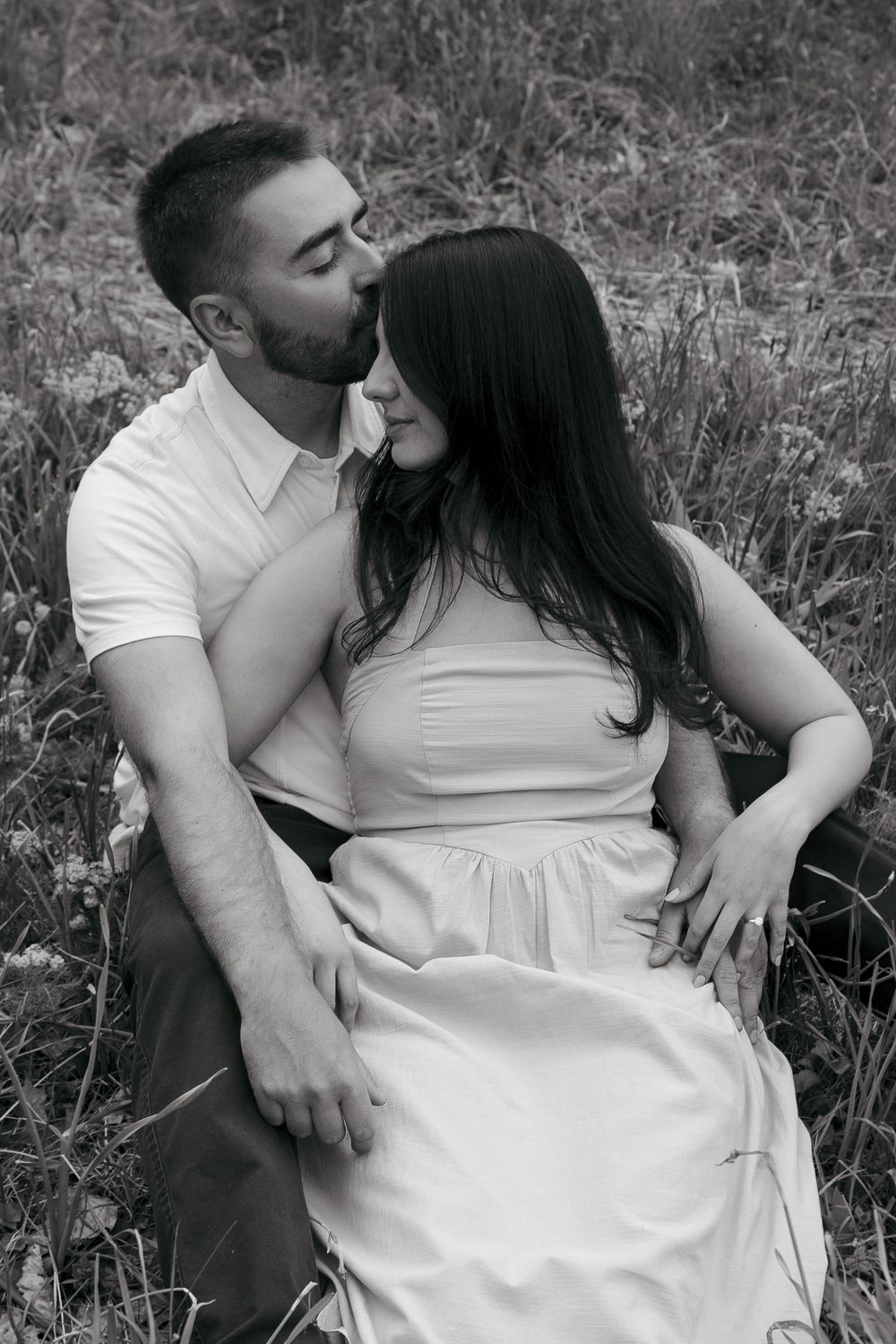 Black and white photo of a couple sitting in a field, man kissing woman on the cheek while embracing her tenderly.