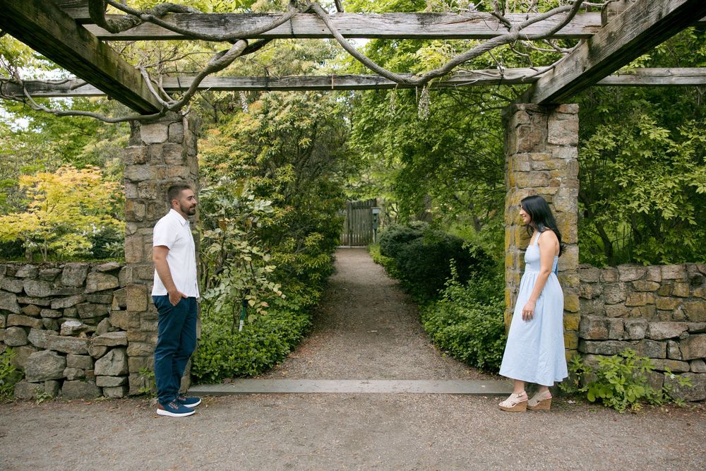 Couple facing each other under a stone pergola archway surrounded by lush green garden foliage.
