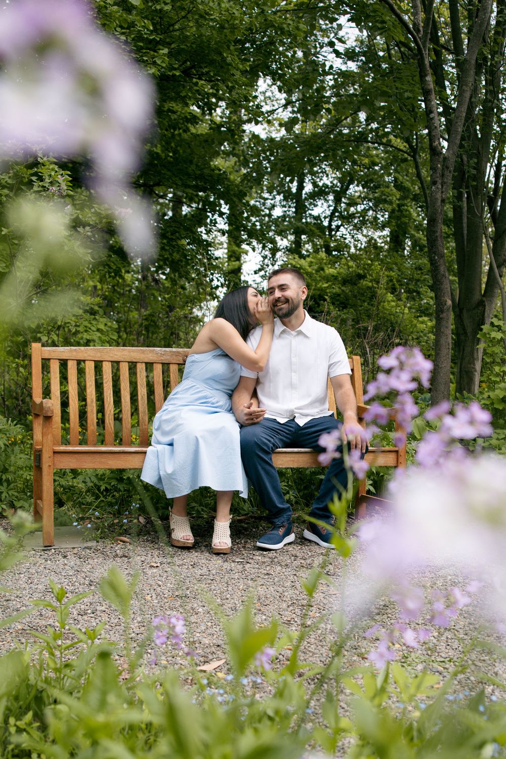 Couple sitting on a wooden bench in a lush garden, woman in blue dress leaning toward smiling man in white shirt.