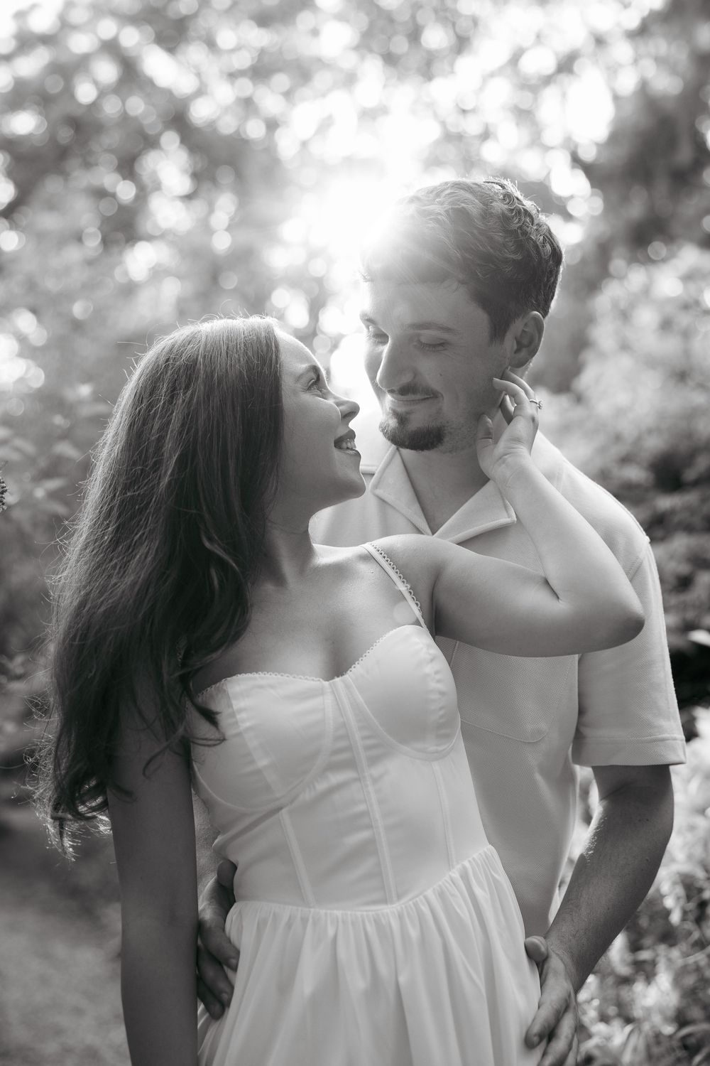 Black and white photo of a romantic couple gazing at each other outdoors, woman in white dress touching mans face.