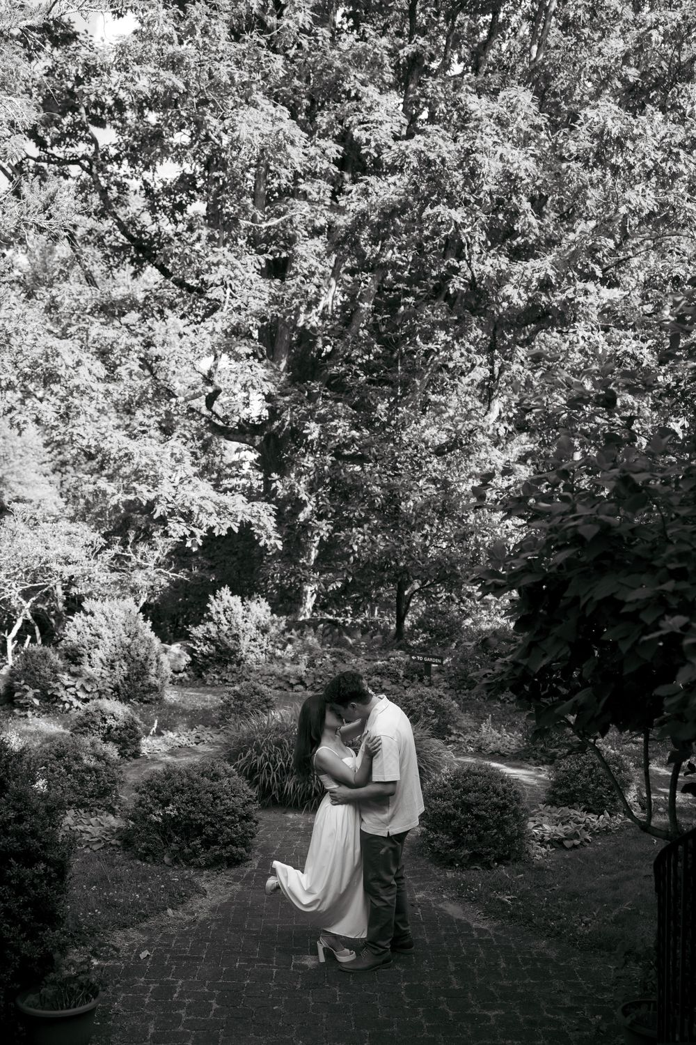Black and white photo of a couple embracing and kissing in a lush garden pathway surrounded by trees.