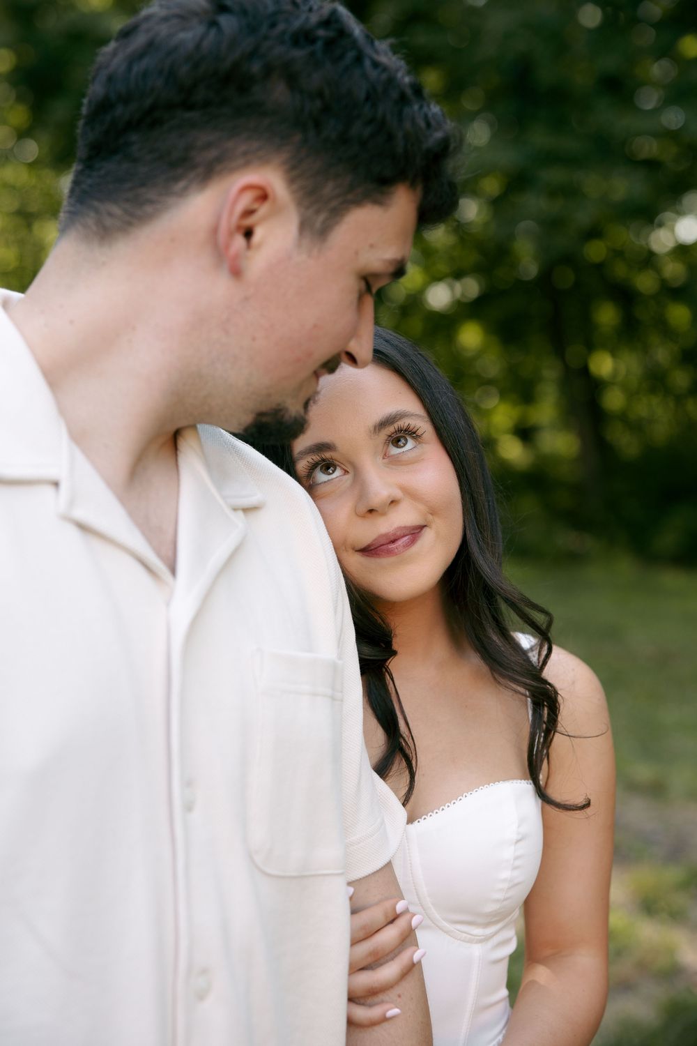 A bride in a white strapless dress leans against a groom in a white shirt outdoors among green trees.