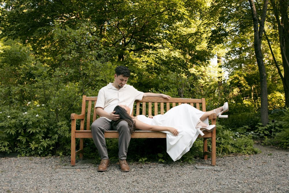 Couple relaxing on a wooden bench in a lush garden, man reading a book while woman lies with legs up.
