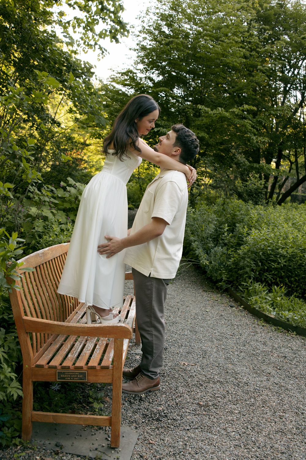 Couple sharing a romantic moment in a lush garden, woman in white dress standing on bench, man tenderly holding her.