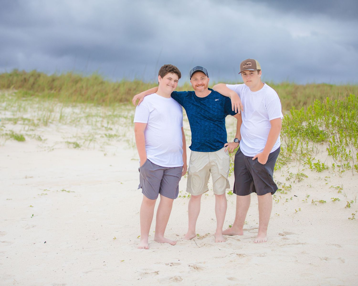 Three friends stand together on a sandy beach with storm clouds gathering overhead.