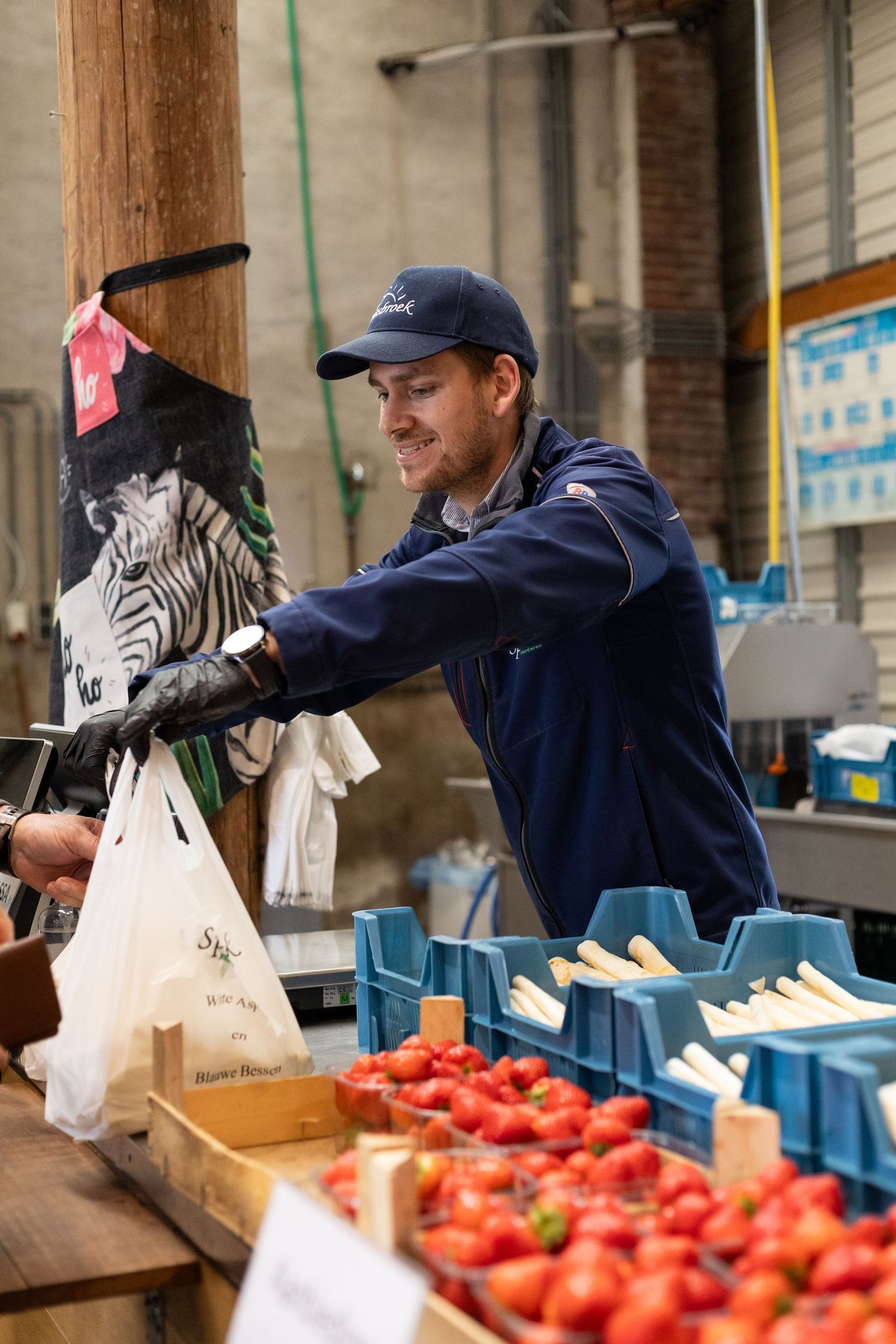 Worker in baseball cap handling fresh tomatoes and produce at a market counter with shopping bags visible.