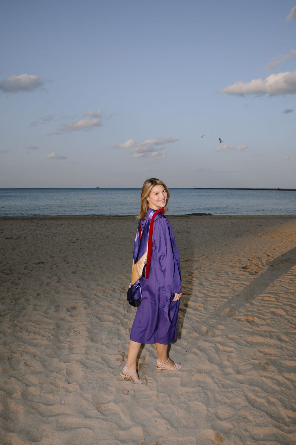 A person wearing a purple jacket and white dress walks along the beach with the ocean in the background.
