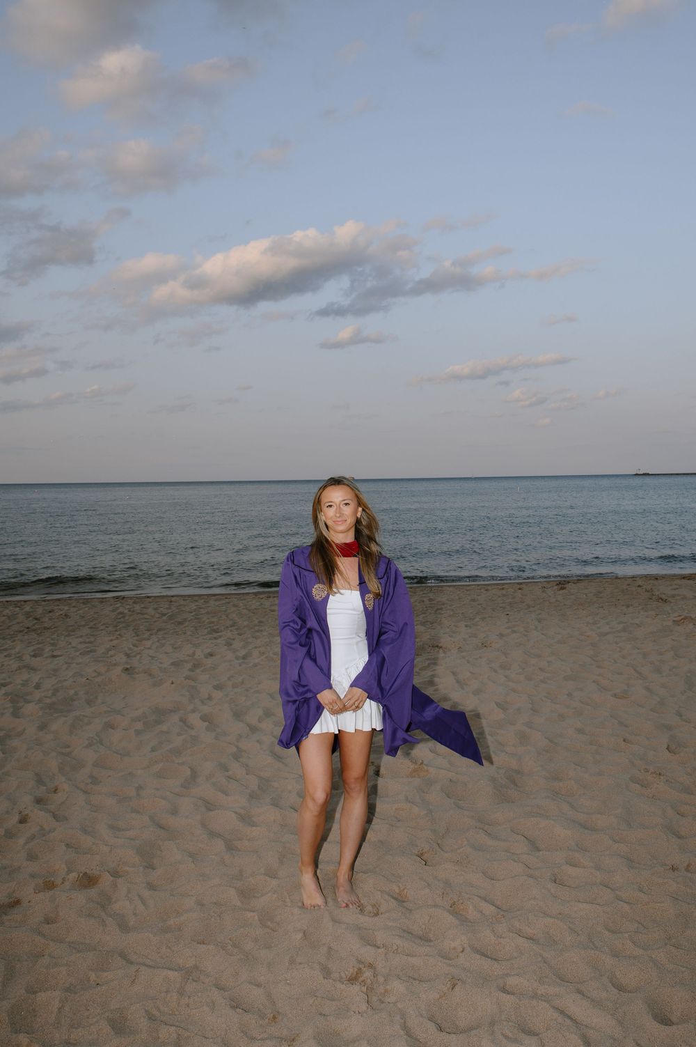 Someone wearing a purple jacket and white shorts stands barefoot on a sandy beach at dusk with ocean and clouds in background.