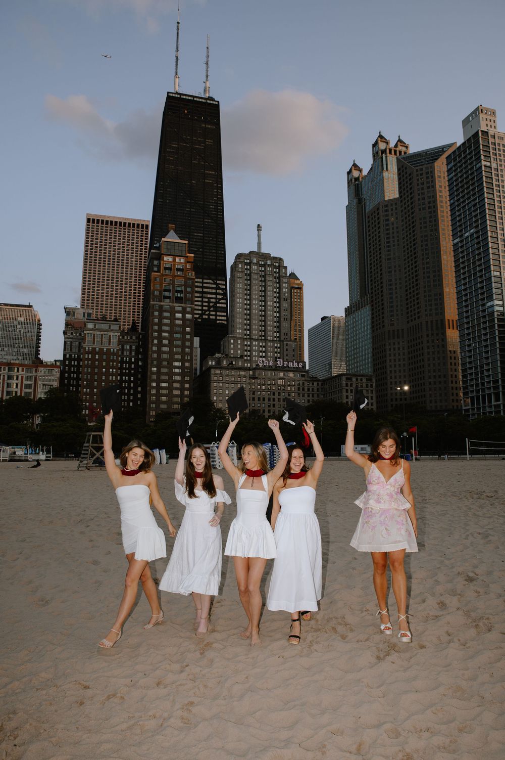 A group in white dresses poses together on a beach with the Chicago skyline behind them.