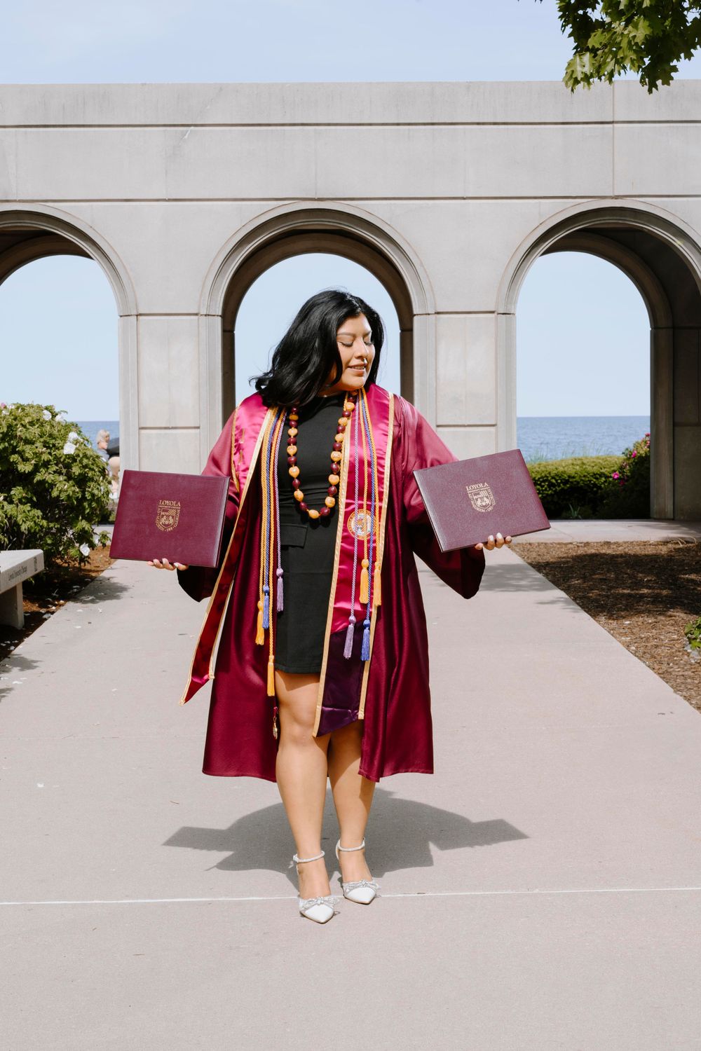 Graduate in maroon gown with diplomas by the lake. Chicago graduation photographer capturing proud moments