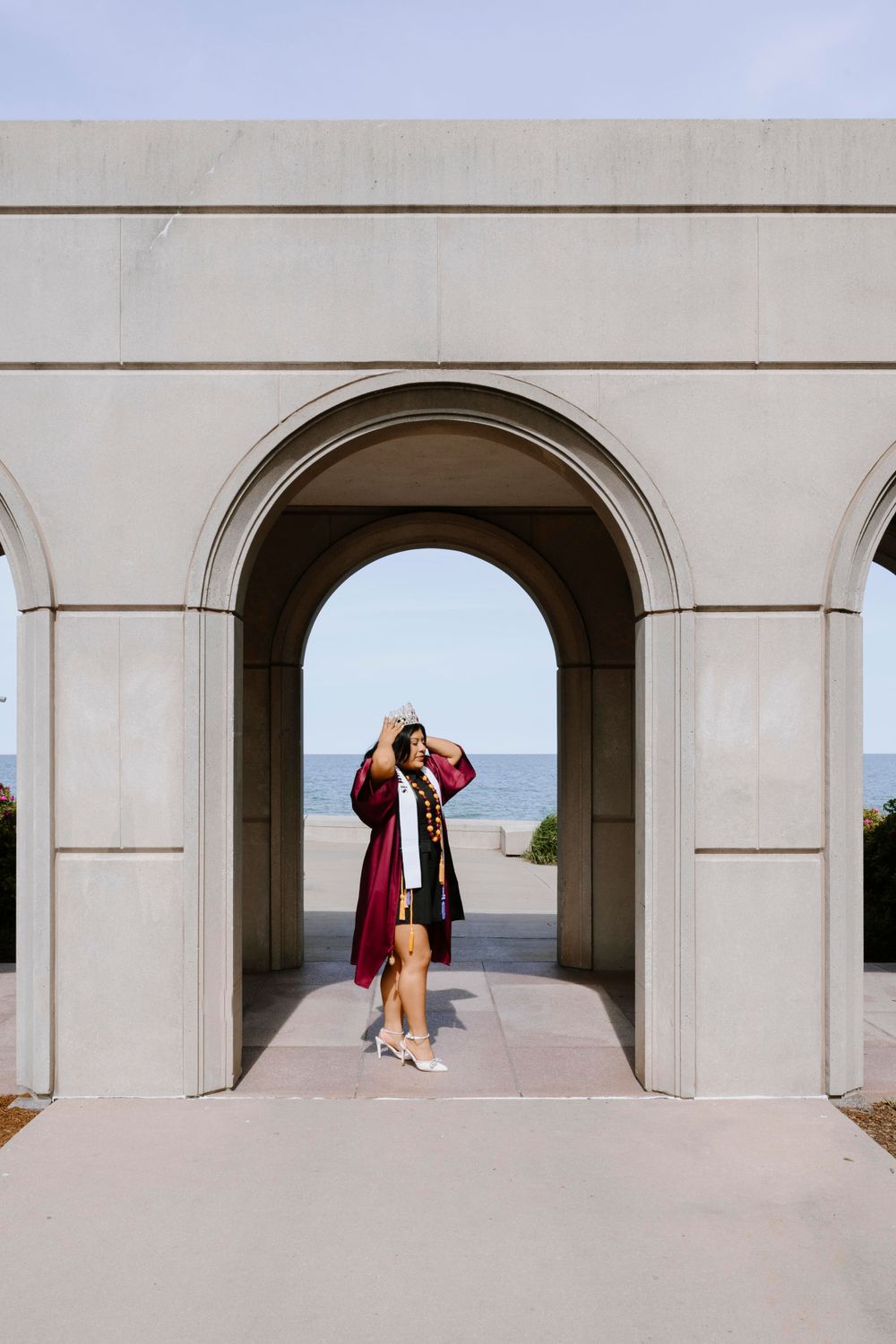 Graduate in maroon gown posing under an archway by the lake. Chicago graduation photographer capturing proud moments