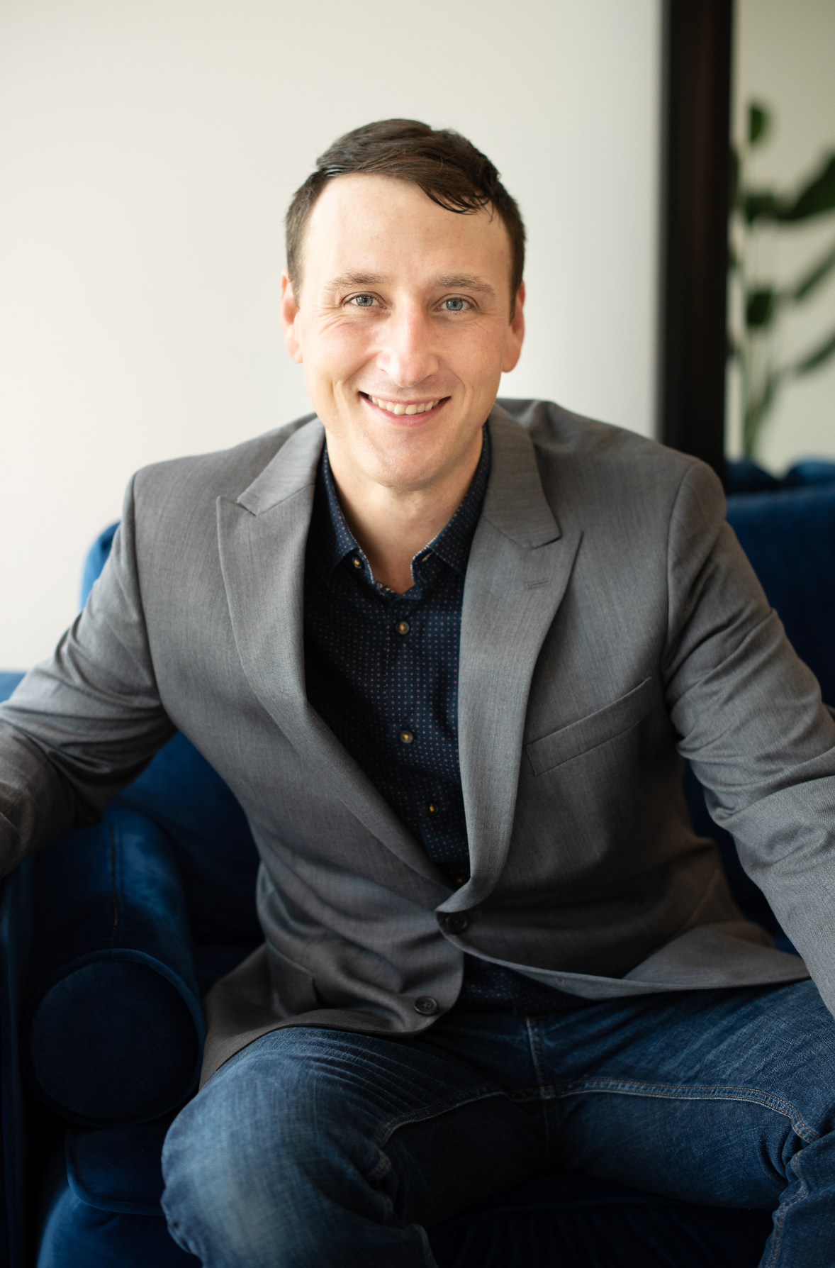 Professional headshot of person in grey blazer and dark shirt seated on blue couch.