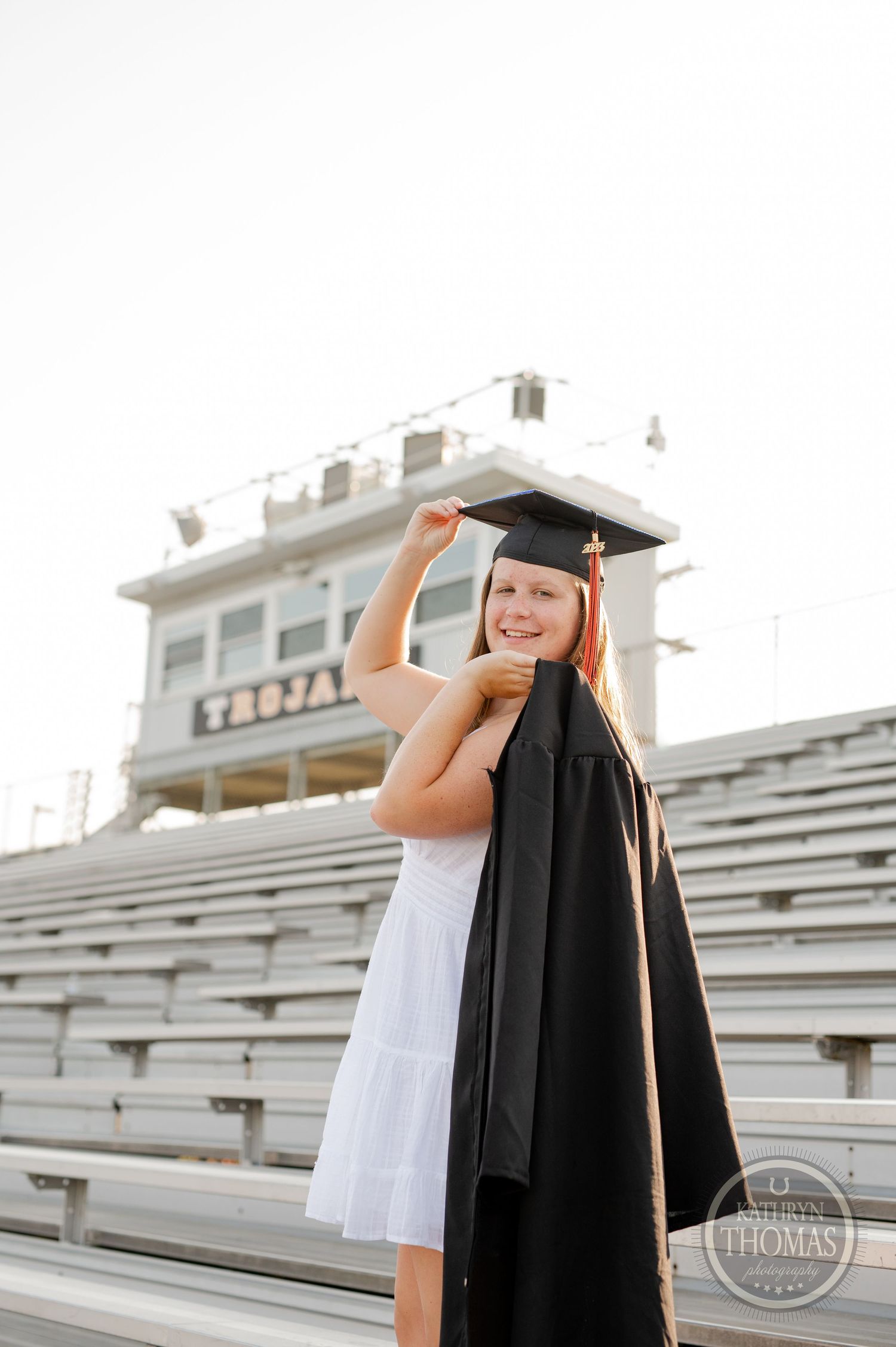 Graduate in white dress and black cap and gown poses on stadium bleachers for graduation photos.
