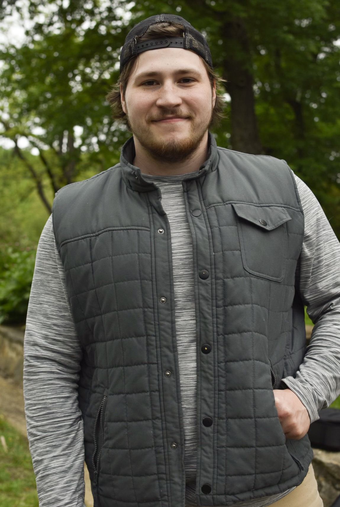 Person wearing a black quilted vest and gray striped sweater poses outdoors among trees.