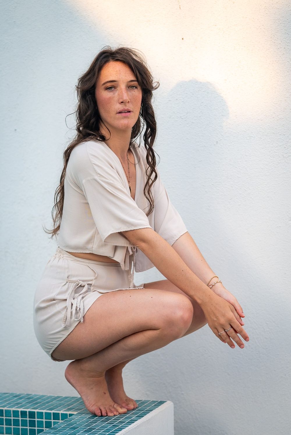 Person in cream colored outfit crouching by pool edge against white wall.
