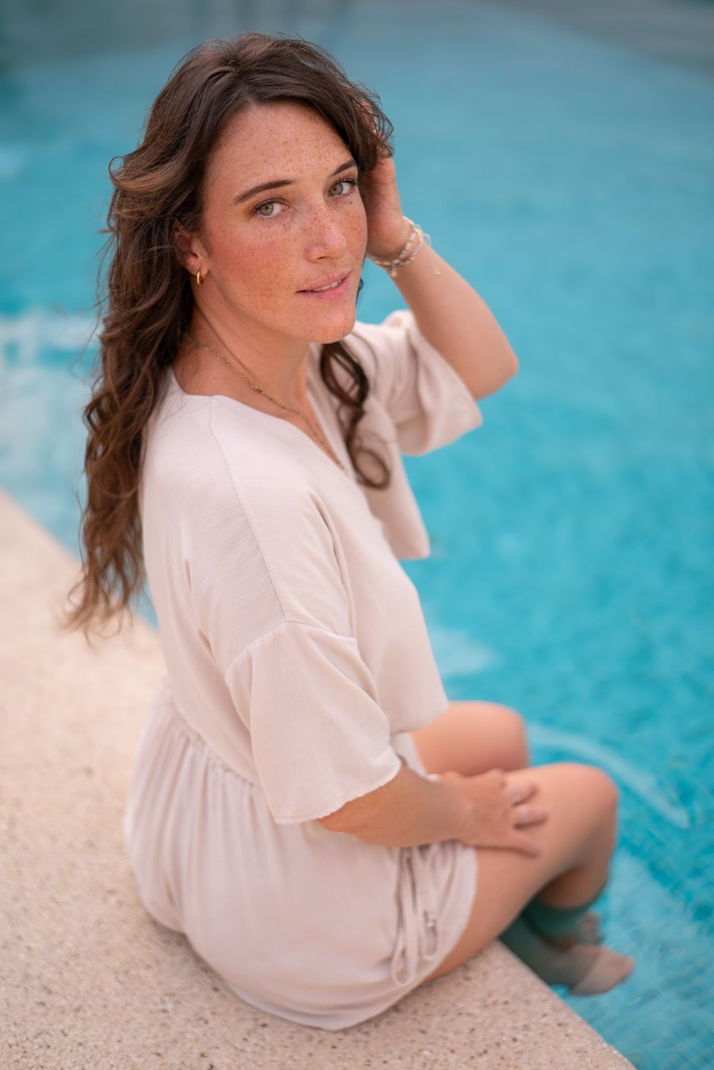Person in cream colored dress sitting poolside with turquoise water background.